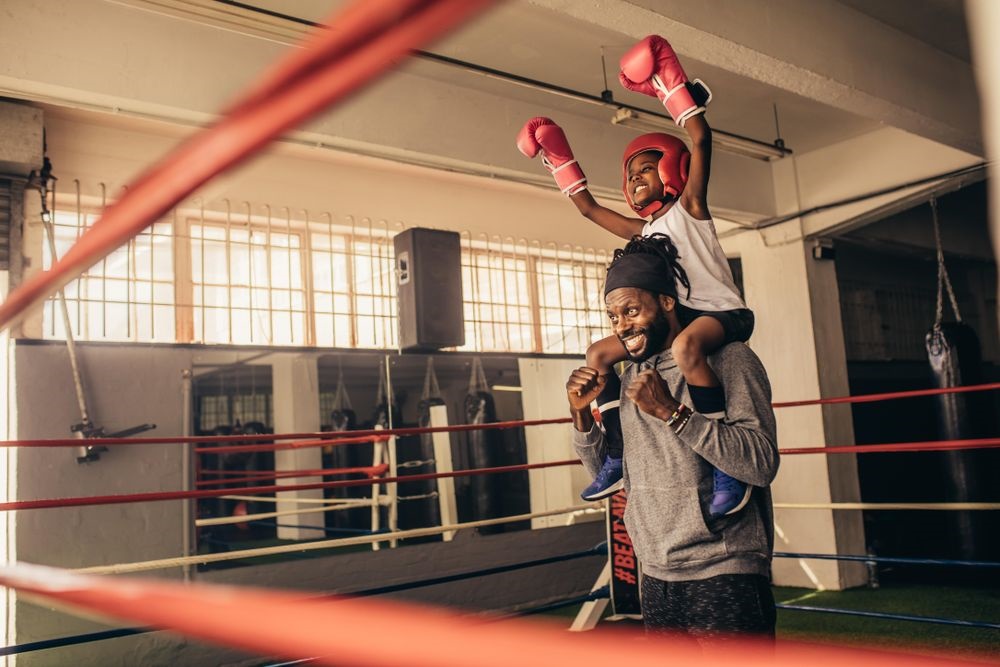 A girl and her father are at a boxing ring. She's siting on his shoulders, smiling triumphantly